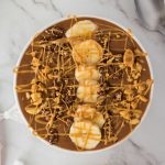Chocolate peanut butter smoothie bowl on a table with sliced bananas, peanuts, peanut butter drizzle and chocolate on top of the smoothie bowl.
