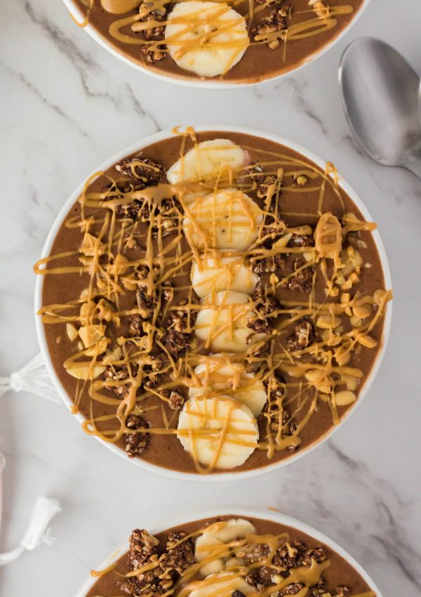 Chocolate peanut butter smoothie bowl on a table with sliced bananas, peanuts, peanut butter drizzle and chocolate on top of the smoothie bowl.