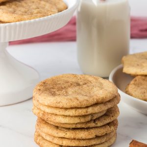 A stack of vegan snickerdoodle cookies on a counter with a glass of vegan milk behind it.