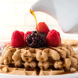 Vegan waffles stacked on a plate with berries on top being drizzled with maple syrup on top.