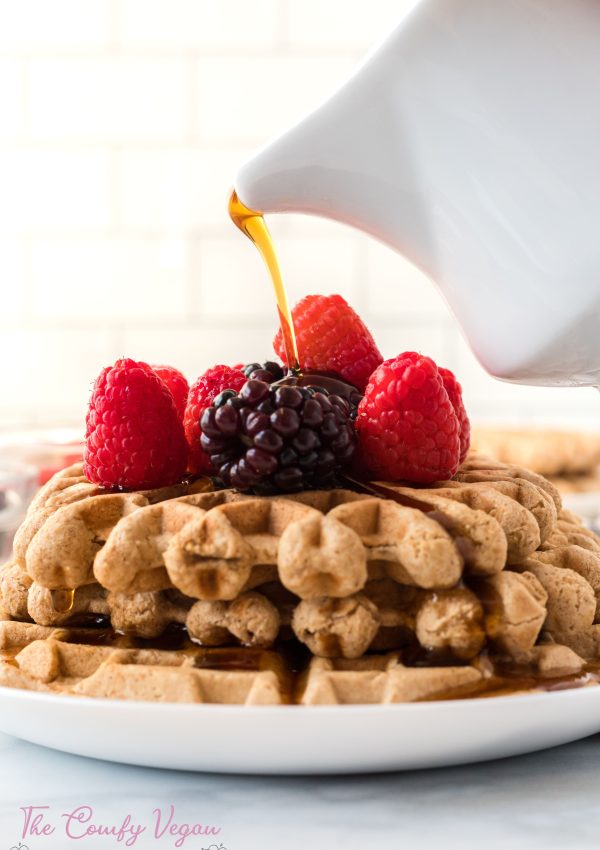 Vegan waffles stacked on a plate with berries on top being drizzled with maple syrup on top.